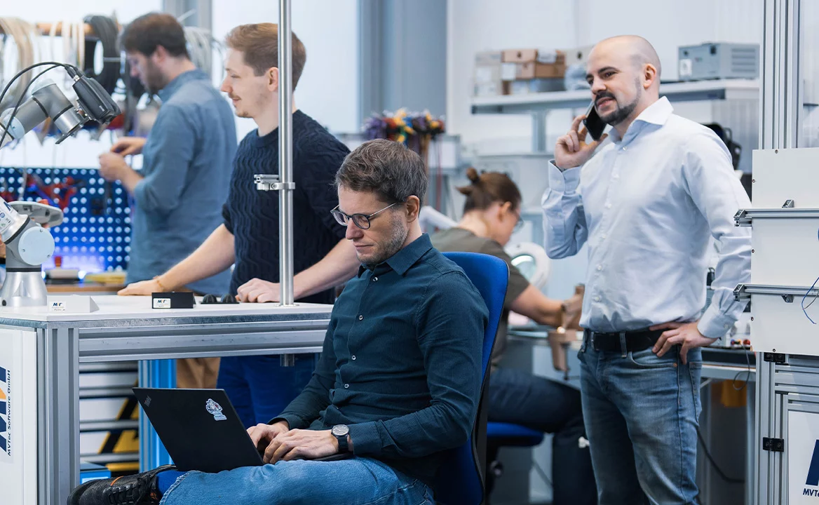 [Translate to Chinese:] Support team in an engineering lab: one person works on a laptop while others test a robotic arm and hardware at a workbench; a colleague is on the phone.