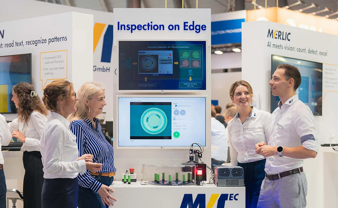 Visitors and MVTec staff discuss an “Inspection on Edge” machine vision demo at a trade show booth, with screens showing visual inspection results.
