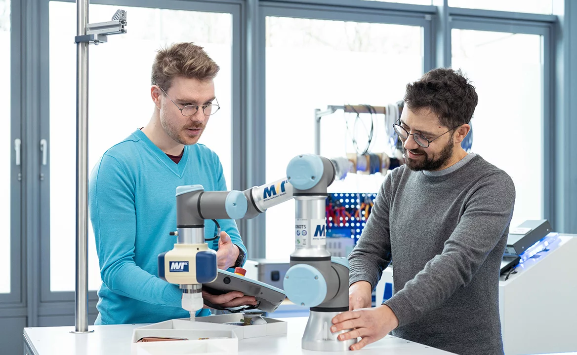 Two engineers set up and operate a collaborative robot arm in a lab environment, using a control panel at a workbench.